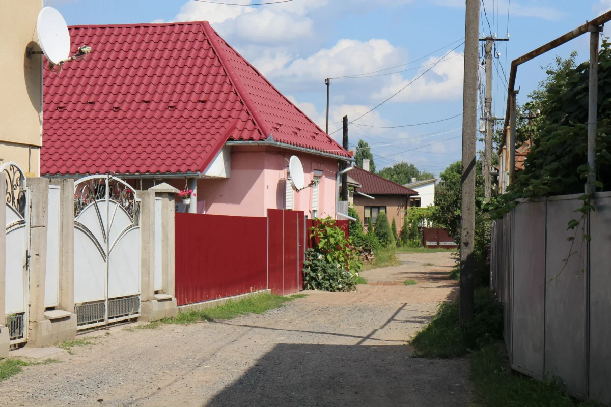 Residential alley with red tiled roofs, satellite dish, and white fences along a sunny street.