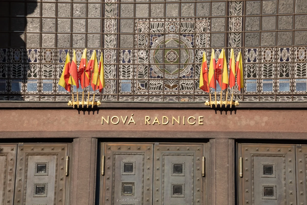 Facade of a historic building with intricate tilework and rows of yellow-red flags in front, reading NOVÁ RADNICE beneath.