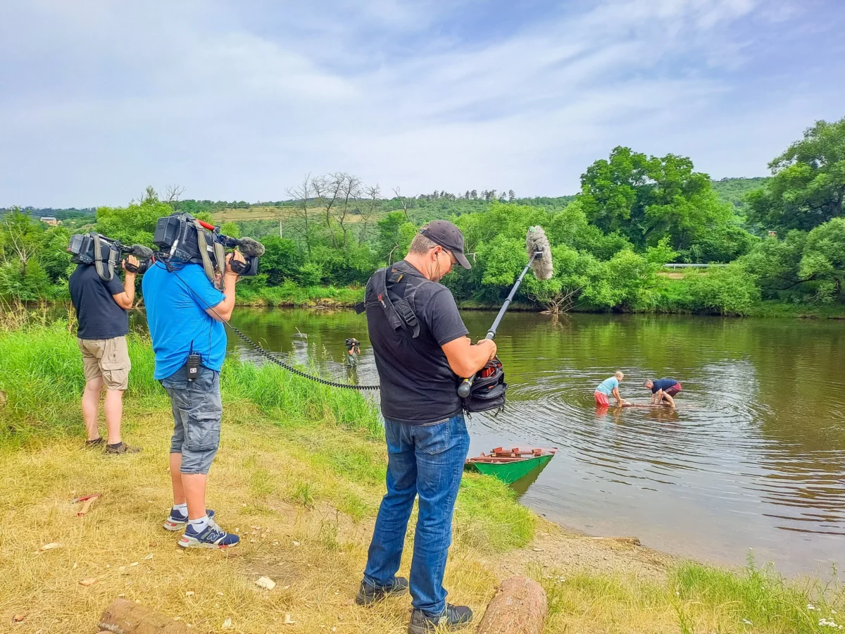 News crew filming at a riverbank with oversized cameras and a boom mic nearby.