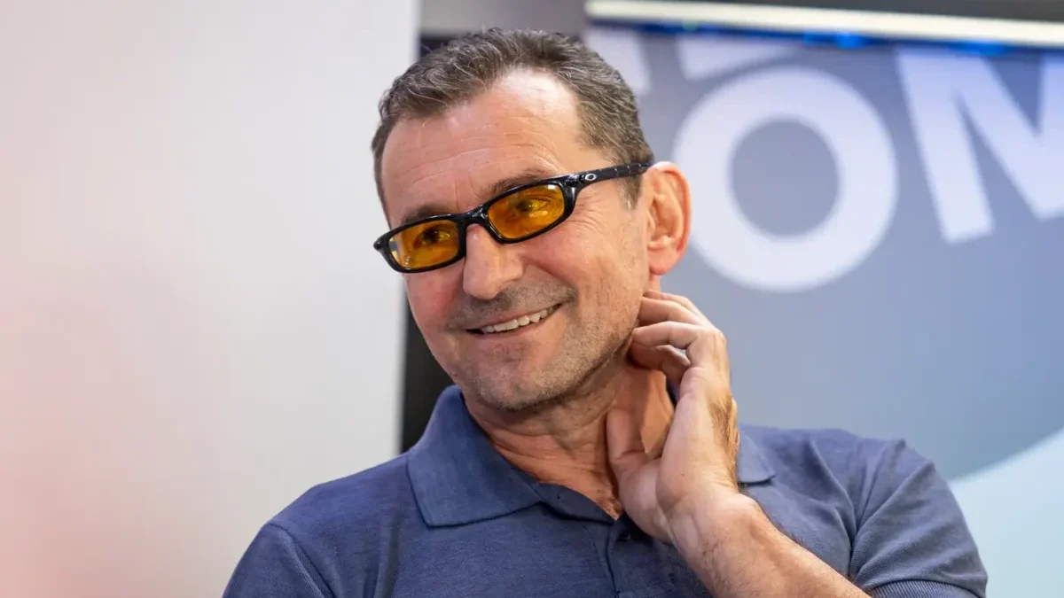 Man smiling, wearing a blue polo shirt and orange-tinted glasses, touching his neck.