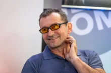 Man smiling, wearing a blue polo shirt and orange-tinted glasses, touching his neck.