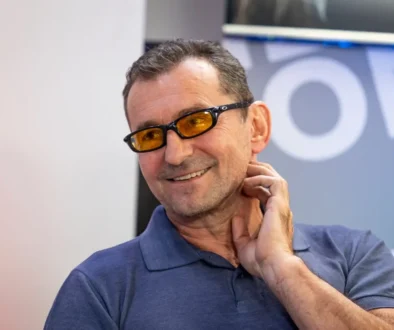 Man smiling, wearing a blue polo shirt and orange-tinted glasses, touching his neck.