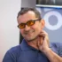 Man smiling, wearing a blue polo shirt and orange-tinted glasses, touching his neck.
