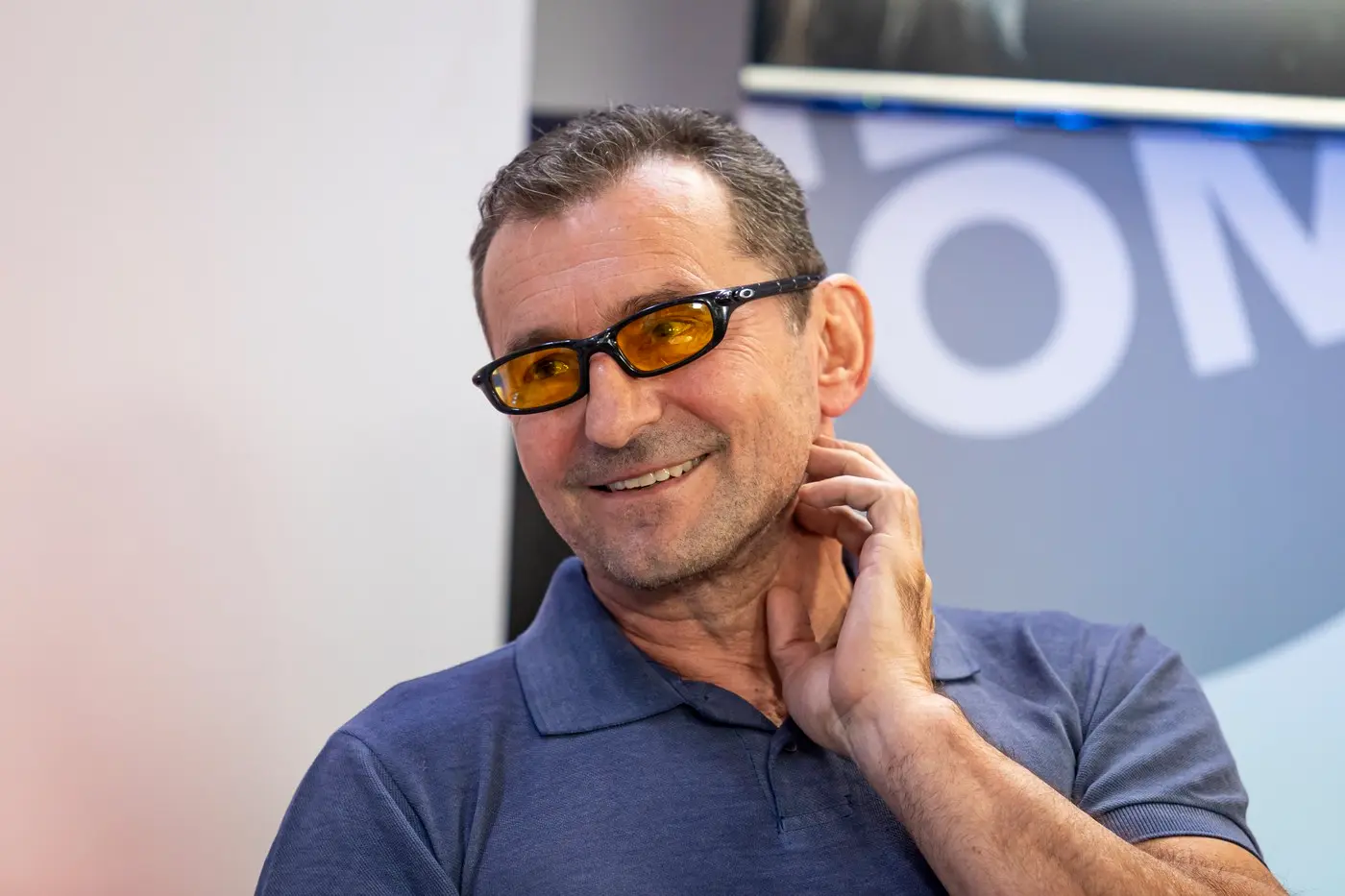 Man smiling, wearing a blue polo shirt and orange-tinted glasses, touching his neck.
