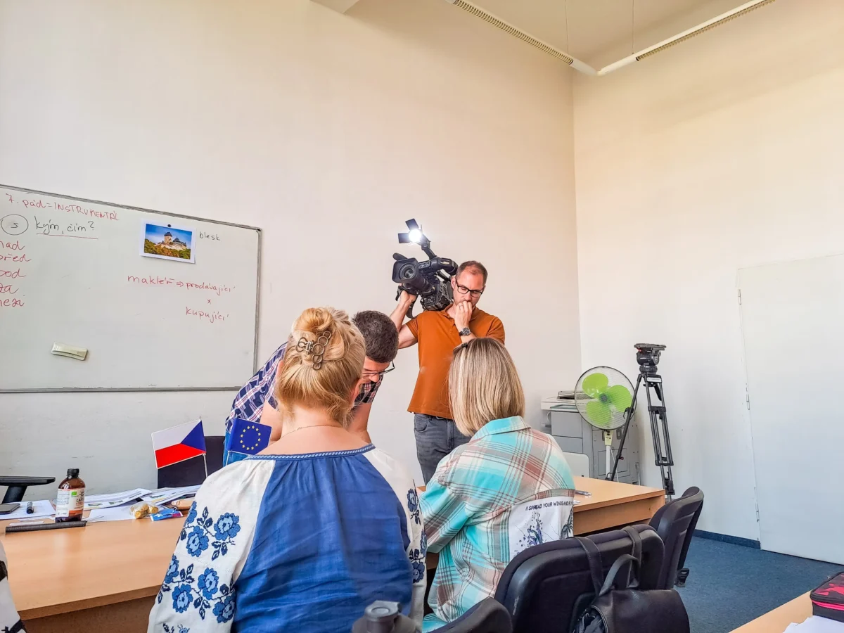 shutterstock_2447919525 1 | TVGURU.cz Group of people seated around a table in a classroom as a man films with a video camera; whiteboard with notes and small flags on the desk are visible.