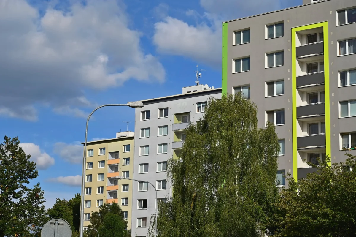 shutterstock_2580856863 1 | TVGURU.cz Modern residential building with bright neon-green vertical trim, grey facade, and white-framed windows, set against a blue sky and clouds; foreground shows a curved street lamp and trees.