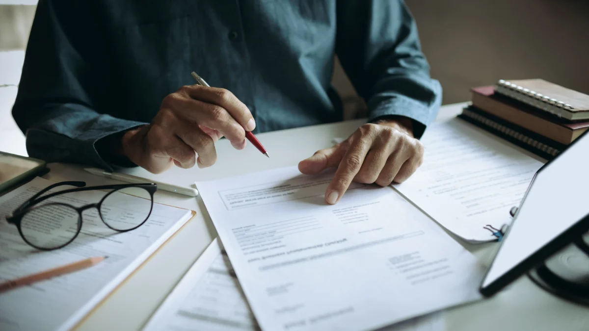 shutterstock_2751810393 1 | TVGURU.cz Person signing papers with a red pen at a cluttered desk, glasses nearby.
