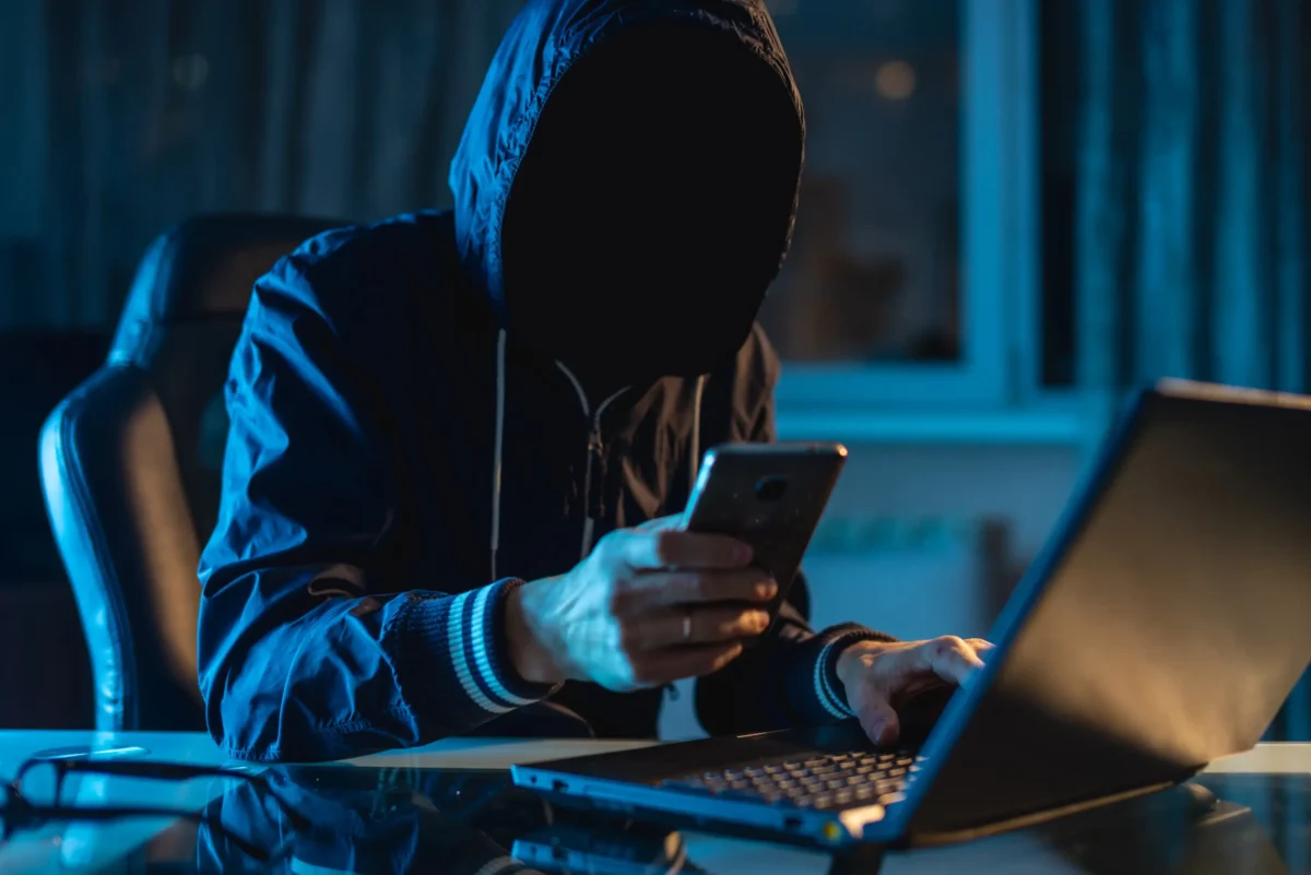 Postava v kápi se dívá na chytrý telefon u stolu s notebookem v tlumeně modře osvětlené místnosti (atmosféra kybernetického hackera).