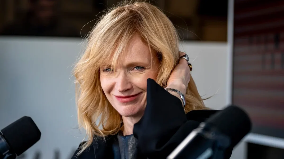 Blonde woman smiling at a microphone during a studio interview, hand touching her hair, wearing a black outfit.