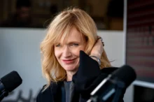 Blonde woman smiling at a microphone during a studio interview, hand touching her hair, wearing a black outfit.