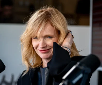 Blonde woman smiling at a microphone during a studio interview, hand touching her hair, wearing a black outfit.