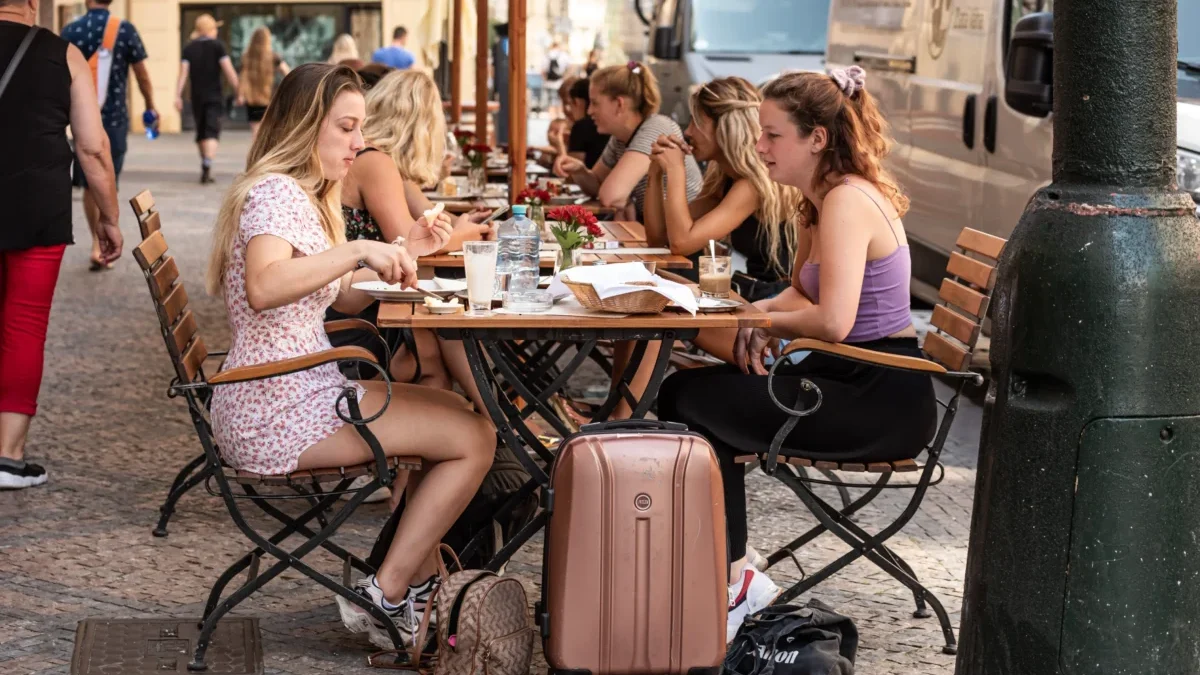 Group of young women dining at an outdoor cafe on a cobblestone street, with suitcases and bags at their feet.