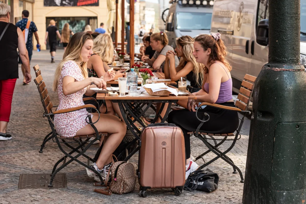 Group of young women dining at an outdoor cafe on a cobblestone street, with suitcases and bags at their feet.
