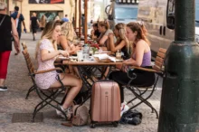 Group of young women dining at an outdoor cafe on a cobblestone street, with suitcases and bags at their feet.