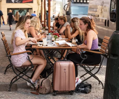 Group of young women dining at an outdoor cafe on a cobblestone street, with suitcases and bags at their feet.