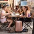 Group of young women dining at an outdoor cafe on a cobblestone street, with suitcases and bags at their feet.