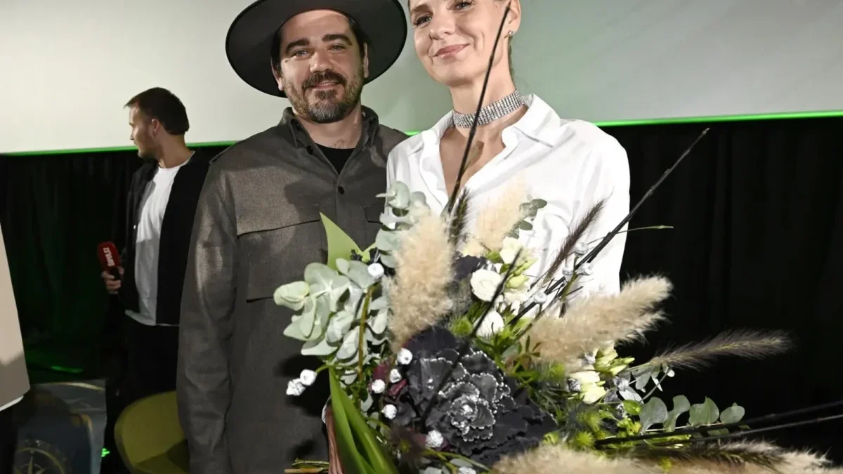 Two people stand on a stage, smiling, with a large bouquet of white and green flowers in the foreground.
