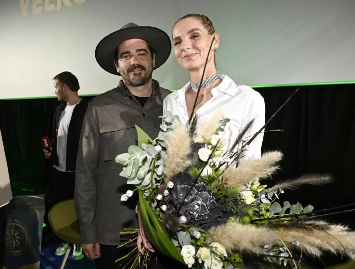 Two people stand on a stage, smiling, with a large bouquet of white and green flowers in the foreground.