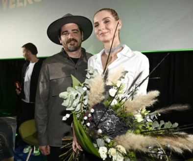 Two people stand on a stage, smiling, with a large bouquet of white and green flowers in the foreground.