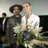 Two people stand on a stage, smiling, with a large bouquet of white and green flowers in the foreground.