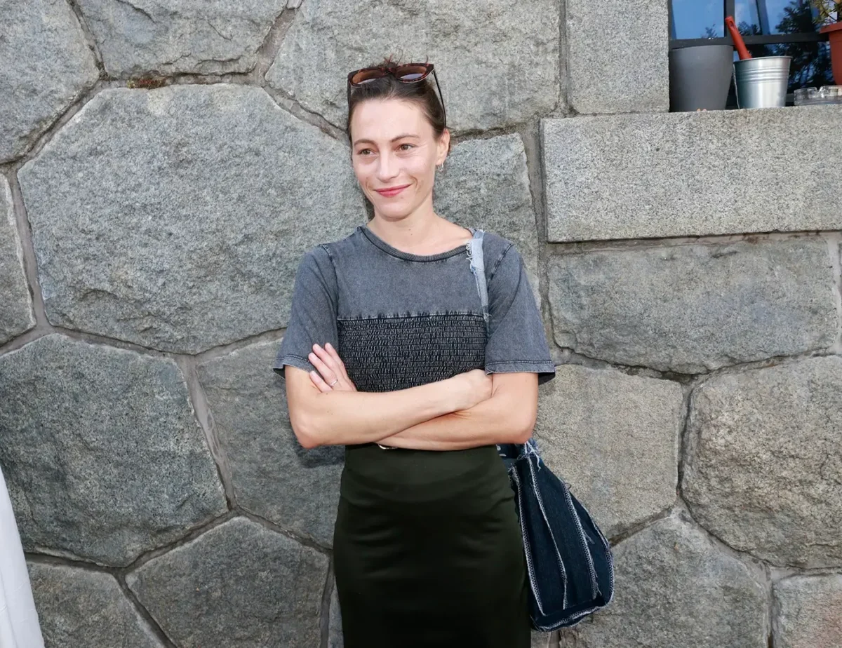 Žena v tmavém oblečení a s tmavými vlasy stojí u kamenné zdi