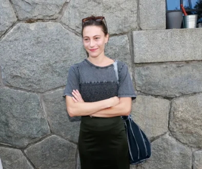 Žena v tmavém oblečení a s tmavými vlasy stojí u kamenné zdi
