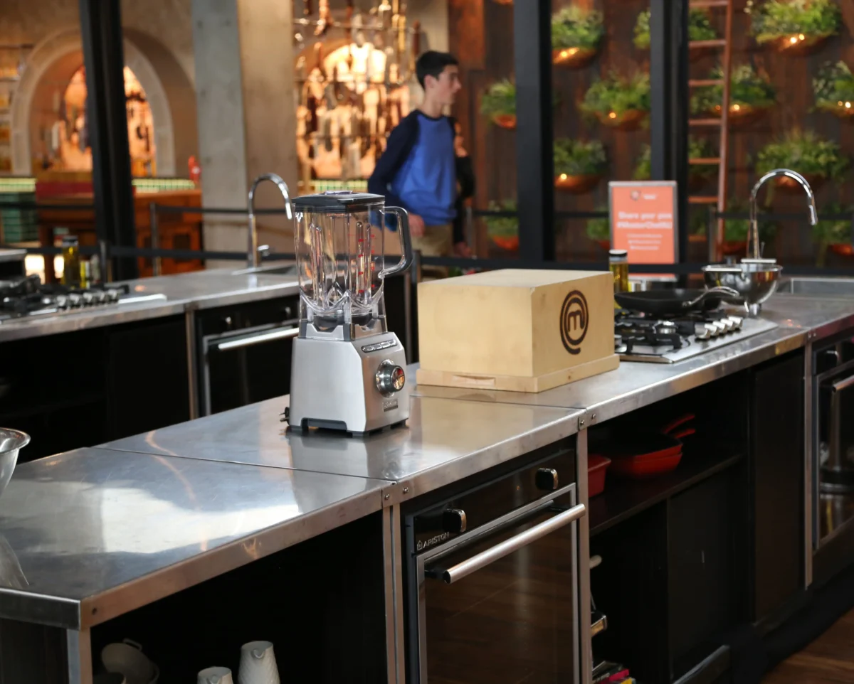 Blender on a stainless steel counter in a modern kitchen studio, with a blurred person in a blue shirt in the background and shelves of equipment behind them.