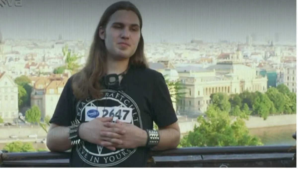 superstar 1 | TVGURU.cz Person with long hair wearing a black graphic T-shirt and race bib 2647, standing on a railing with a city skyline behind.