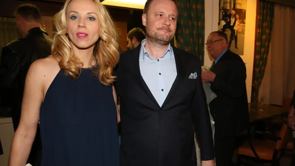 Blonde woman in a navy dress stands beside a man in a dark suit at an indoor event, warm lighting in the background.