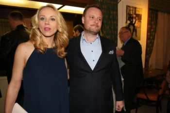 Blonde woman in a navy dress stands beside a man in a dark suit at an indoor event, warm lighting in the background.