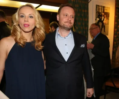 Blonde woman in a navy dress stands beside a man in a dark suit at an indoor event, warm lighting in the background.