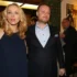 Blonde woman in a navy dress stands beside a man in a dark suit at an indoor event, warm lighting in the background.