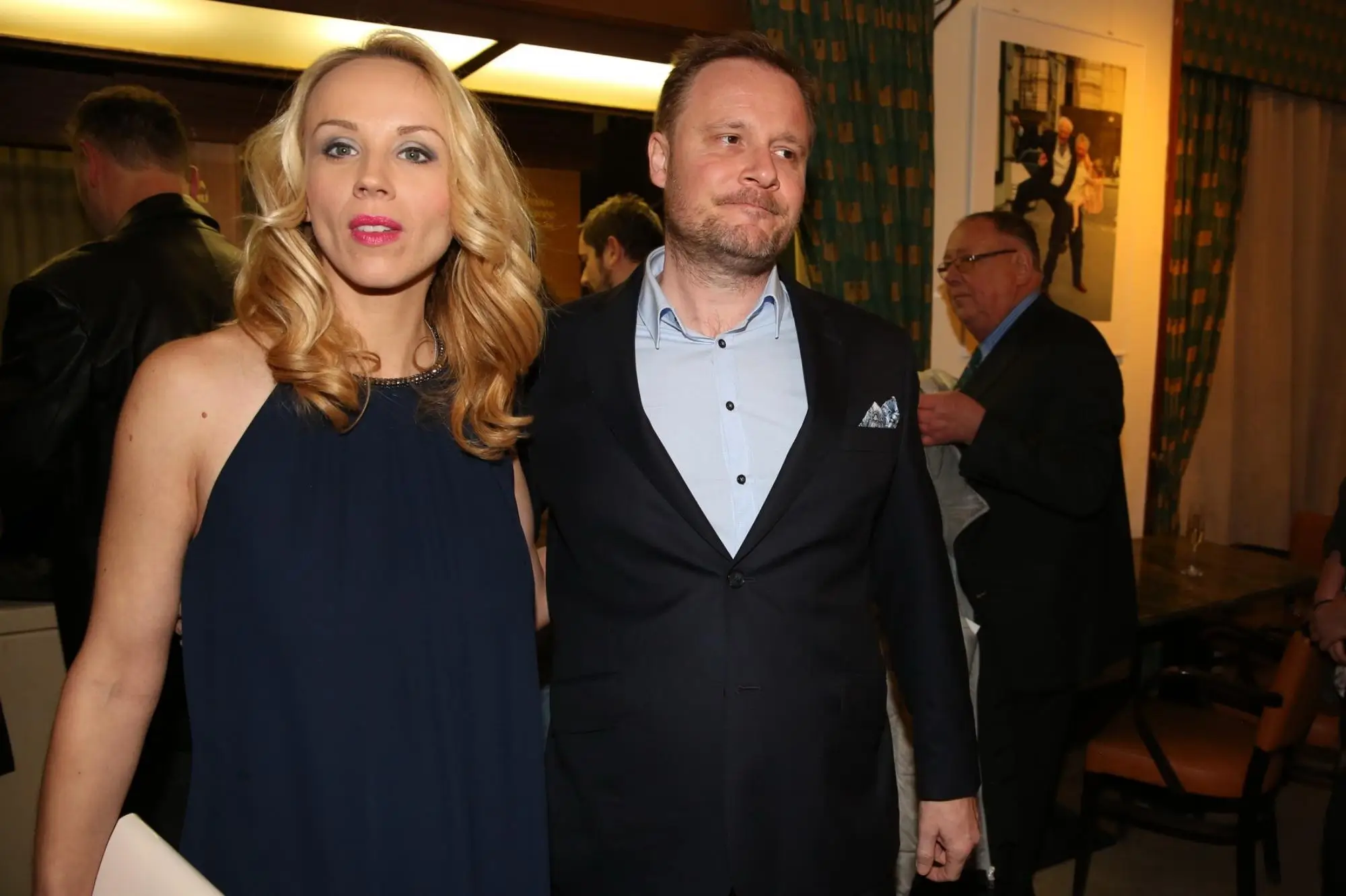 Blonde woman in a navy dress stands beside a man in a dark suit at an indoor event, warm lighting in the background.