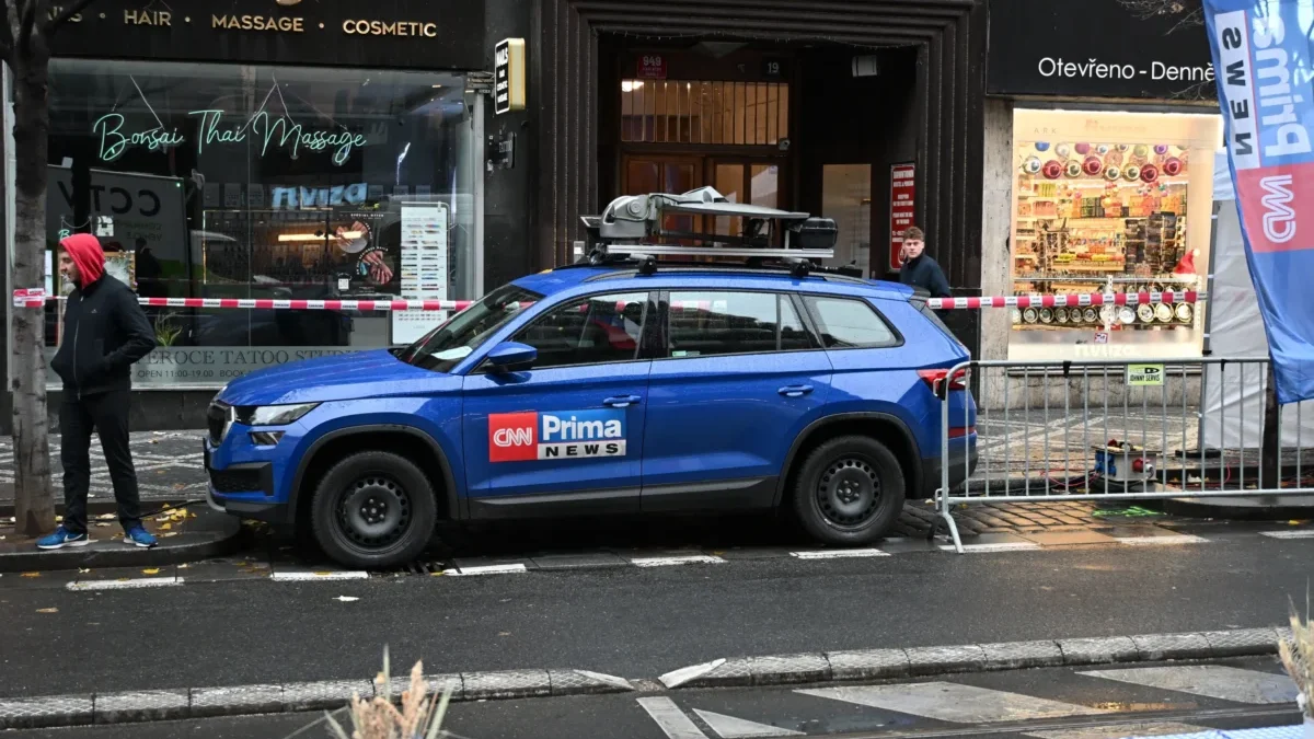 Blue CNN News SUV with roof rack parked on a city street in front of shops; a man in a red hoodie walks along the sidewalk behind caution tape.