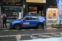 Blue CNN News SUV with roof rack parked on a city street in front of shops; a man in a red hoodie walks along the sidewalk behind caution tape.