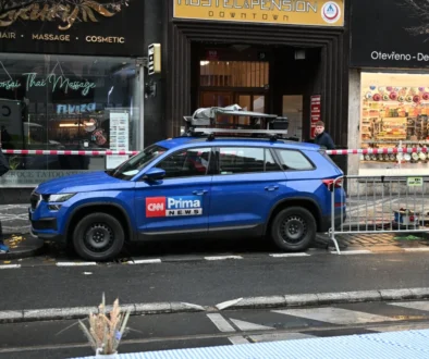 Blue CNN News SUV with roof rack parked on a city street in front of shops; a man in a red hoodie walks along the sidewalk behind caution tape.