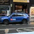Blue CNN News SUV with roof rack parked on a city street in front of shops; a man in a red hoodie walks along the sidewalk behind caution tape.