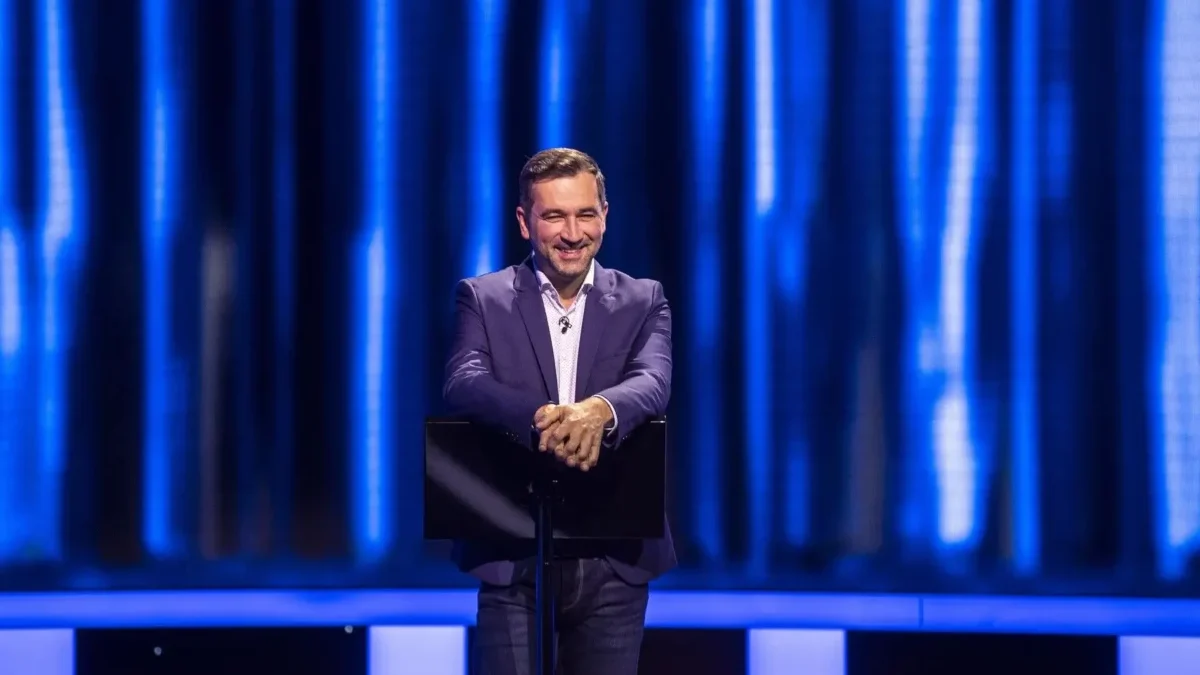 Man in a navy blazer delivering a talk on a stage with a blue curtain backdrop.