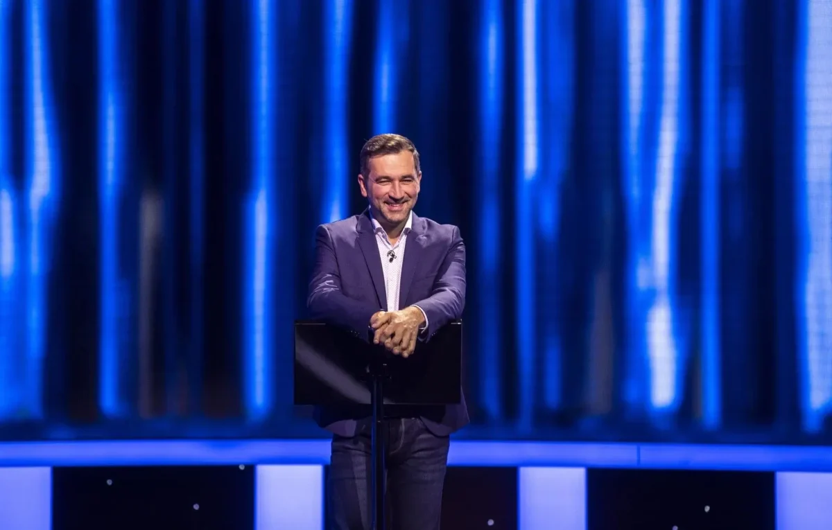 Man in a navy blazer delivering a talk on a stage with a blue curtain backdrop.