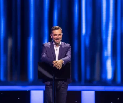 Man in a navy blazer delivering a talk on a stage with a blue curtain backdrop.