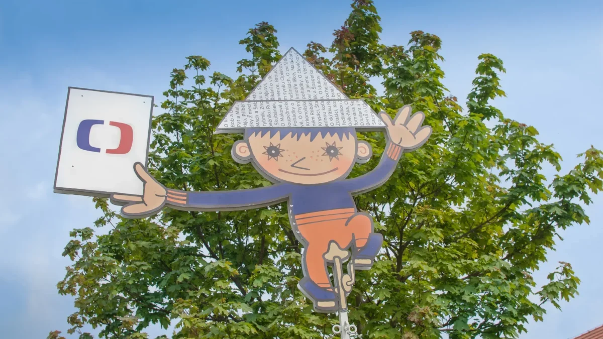 Whimsical cartoon boy cutout hanging from a pole among green trees against a blue sky, with a sign to the left.