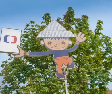 Whimsical cartoon boy cutout hanging from a pole among green trees against a blue sky, with a sign to the left.