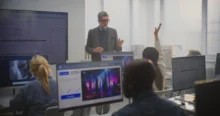 Instructor in a blazer teaches at the front of a computer lab, students raise hands while multiple monitor screens surround them