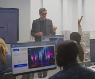Instructor in a blazer teaches at the front of a computer lab, students raise hands while multiple monitor screens surround them