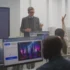Instructor in a blazer teaches at the front of a computer lab, students raise hands while multiple monitor screens surround them