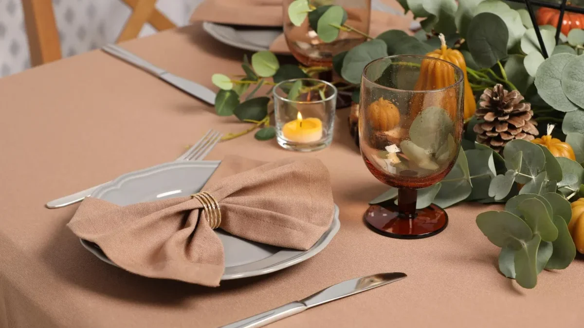Elegant autumn table setting with a folded beige napkin on a plate, polished silverware, an amber wine glass, and a small candle surrounded by greenery and pinecones.