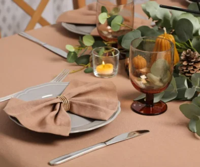 Elegant autumn table setting with a folded beige napkin on a plate, polished silverware, an amber wine glass, and a small candle surrounded by greenery and pinecones.