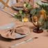 Elegant autumn table setting with a folded beige napkin on a plate, polished silverware, an amber wine glass, and a small candle surrounded by greenery and pinecones.