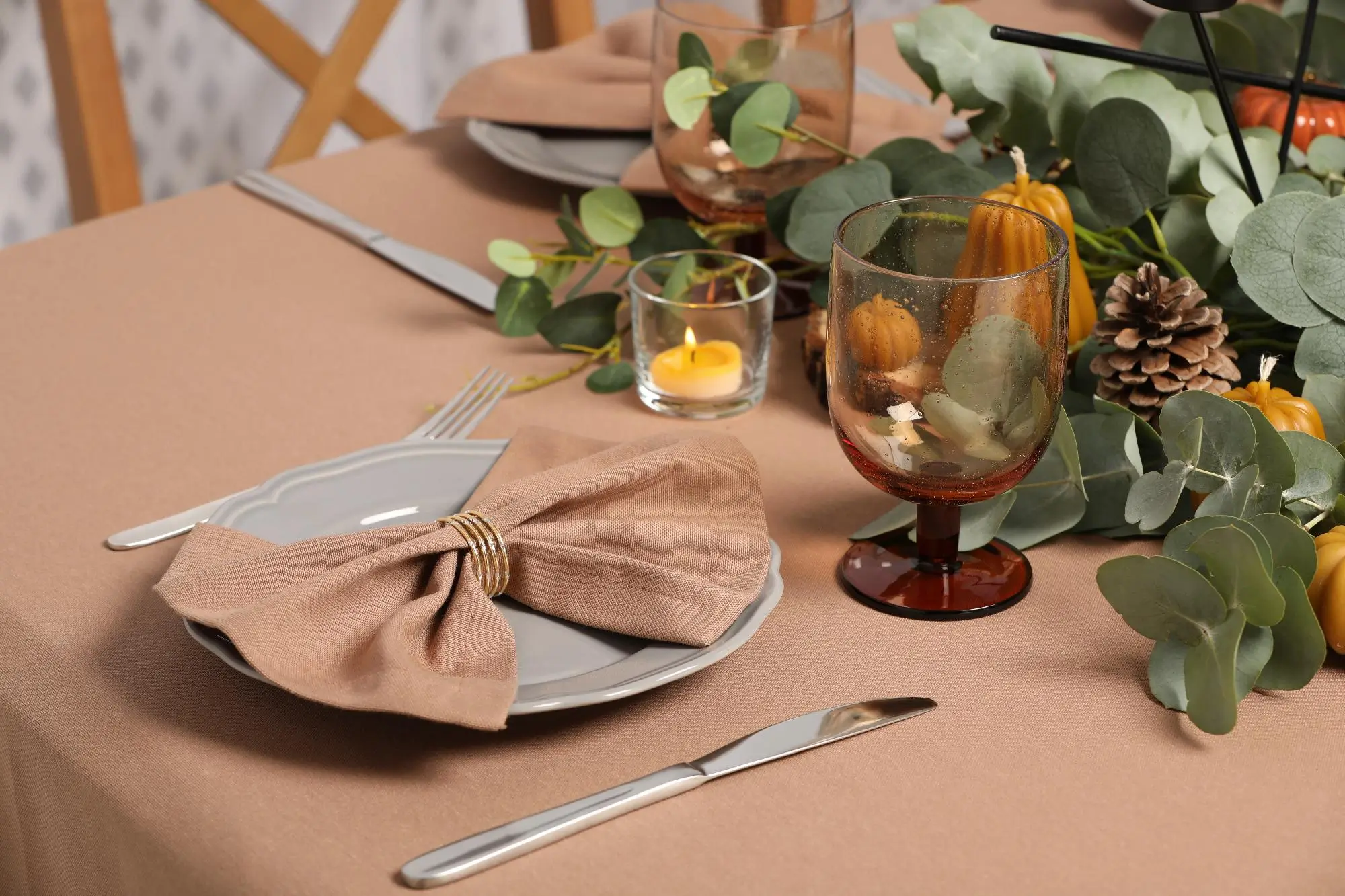 Elegant autumn table setting with a folded beige napkin on a plate, polished silverware, an amber wine glass, and a small candle surrounded by greenery and pinecones.