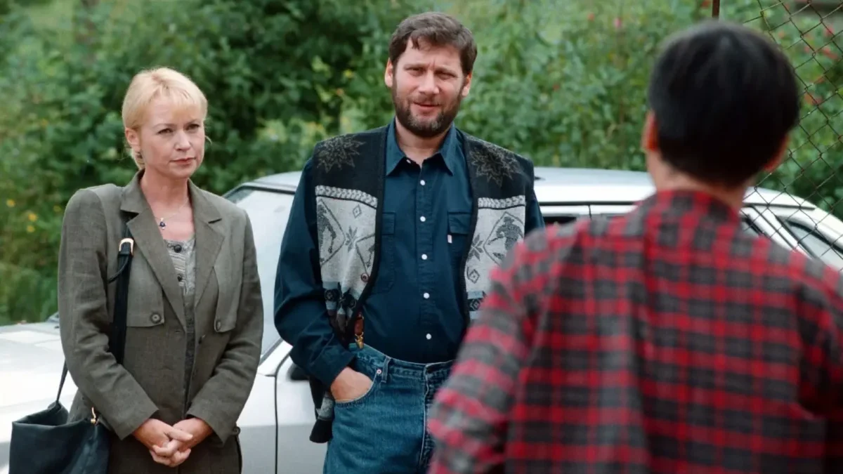Two adults stand by a silver car talking to a person in a red plaid shirt (back to camera) in an outdoor setting with greenery in the background.